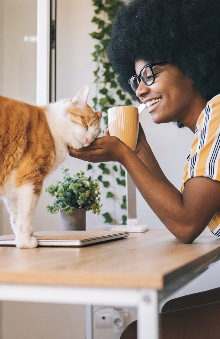 A woman drinking coffee at a desk cradles the head of a happy orange and white cat.
