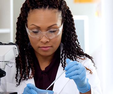Scientist in lab coat wearing gloves and protective glasses using a pipette.