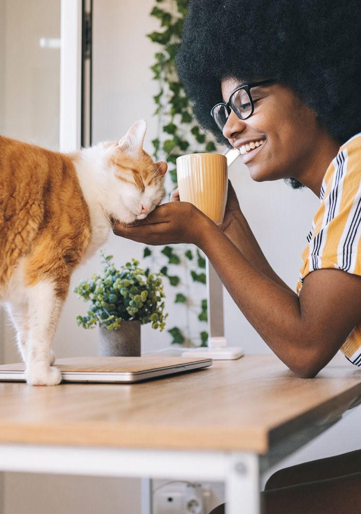 A woman drinking coffee at a desk cradles the head of a happy orange and white cat.