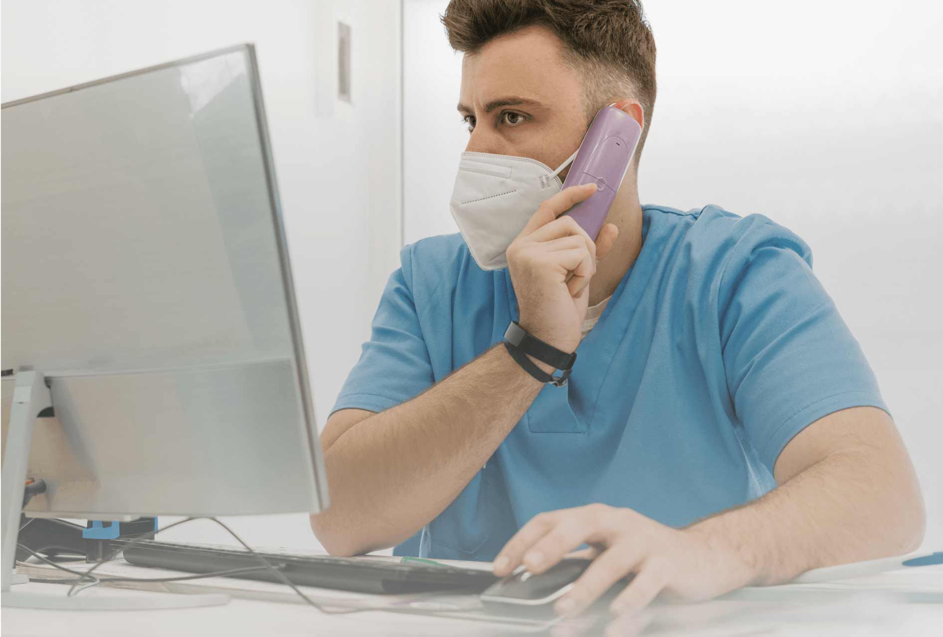 Man in blue scrubs talking on the phone while sitting at a desktop computer.